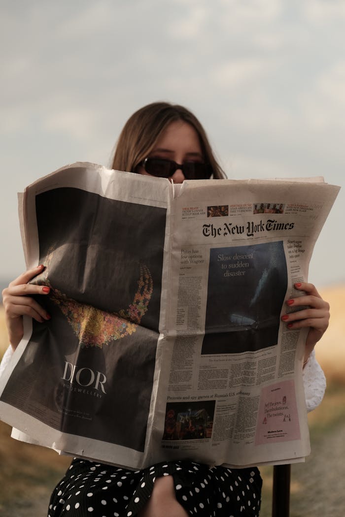 A woman in sunglasses reads The New York Times while seated outside, with a stylish fashion vibe.
